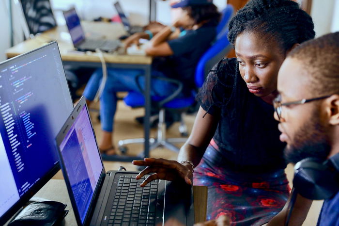Two people work together at a laptop in an office setting, with others working at computers in the background.