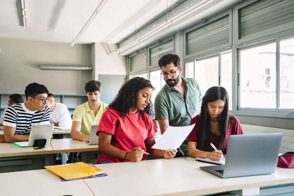 Friendly Professor explaining exercise to students in high school classroom. Young Teacher with beard helping teenager girl - Focus on Male teacher