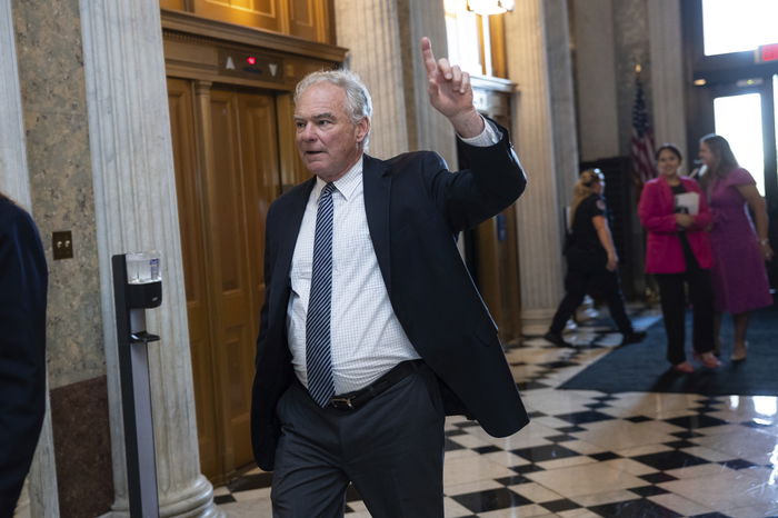 A man in a suit walks through a marble-floored hallway, raising his hand with one finger pointing upward. Other people are visible in the background.