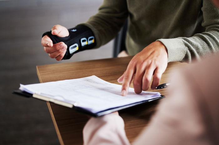 Two people sit at a table reviewing a document. One person wears a wrist brace and points at the paper.