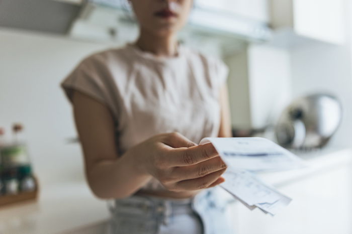 Person extending a few envelopes or papers with one hand, standing in a kitchen environment. Background includes kitchen items on a counter.