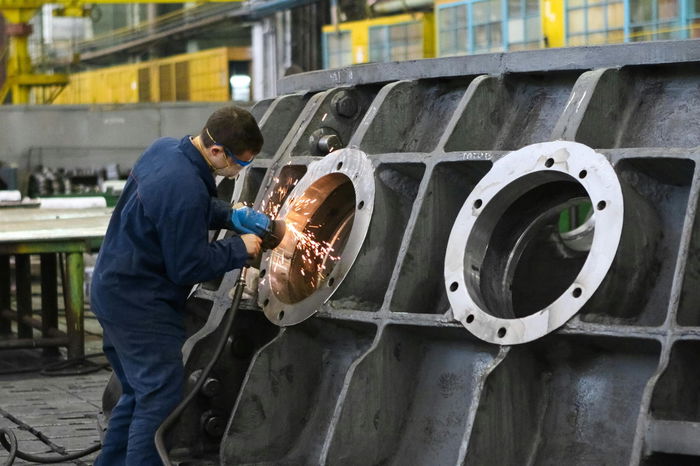 Worker in blue overalls using a power tool to grind a large metal component in an industrial setting.
