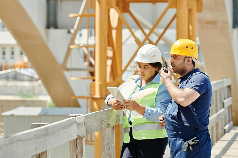 Two construction workers wearing hard hats, one holding a tablet and the other using a walkie-talkie, stand on a wooden bridge at a construction site.