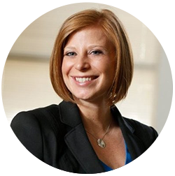 A person with short red hair smiling, wearing a black blazer and a blue top.