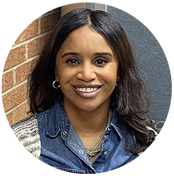 A woman with long dark hair, wearing a denim shirt and hoop earrings, smiles while standing against a brick wall.