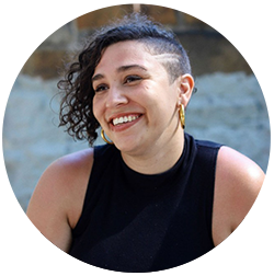 A person with curly hair and an undercut, wearing a black sleeveless top and hoop earrings, smiling against a blurred background.