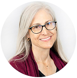 A person with long gray hair and glasses smiles, wearing a dark top and a maroon scarf, against a plain background.