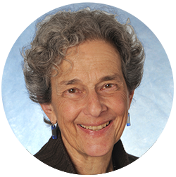A person with curly gray hair and blue earrings smiles against a light blue background.