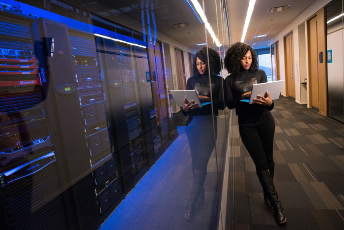 Person with a laptop leans against a glass wall in a server room, surrounded by computer equipment and blue lighting.