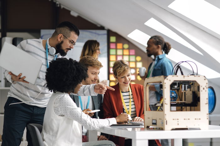 A diverse group of people collaborates around a table with a 3D printer and a laptop in a bright, modern office space.