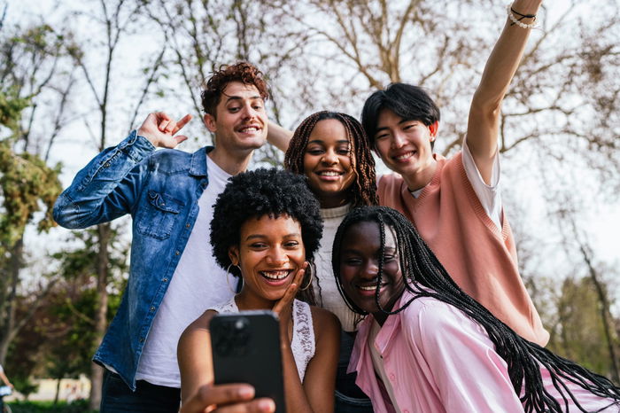 A group of five people stand close together outdoors, smiling and taking a selfie. Trees are visible in the background.