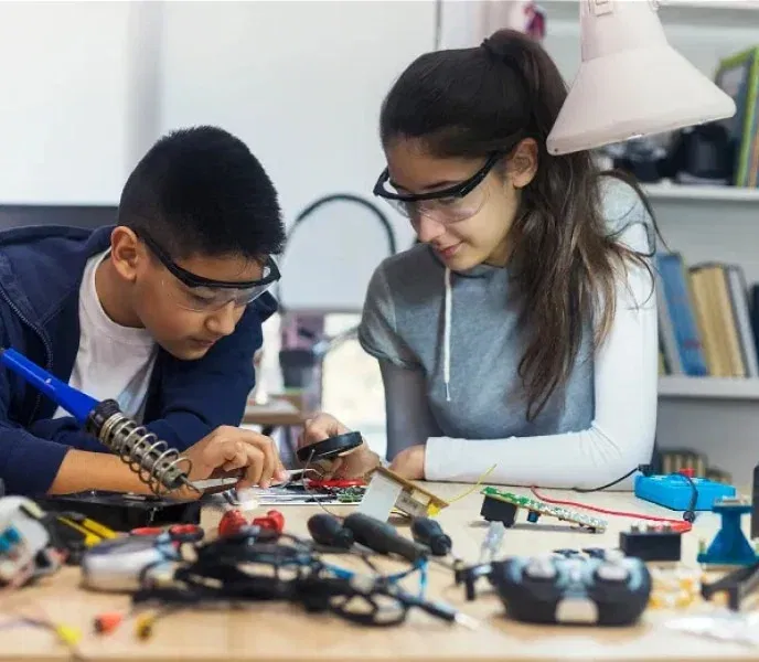 Two people wearing safety goggles work on an electronics project at a table cluttered with tools and components.