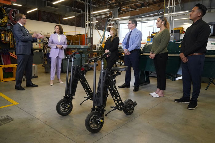 A group of people, including Kamala Harris in a purple suit, are standing in a workshop with two electric scooters in the foreground.