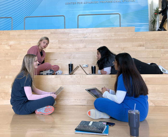 Four female students sitting together in a group on wooden steps, using tablets and laptops, with cups and a notebook nearby.