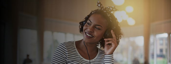 Woman in a striped top, smiling while listening to headphones, with sunlight in the background.