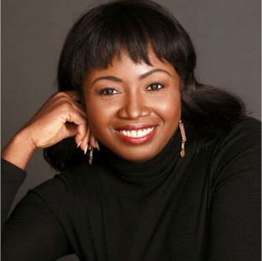 A person smiling, wearing a black turtleneck and earrings, with one hand resting on their chin against a gray background.