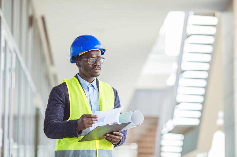 A person in a blue hard hat and yellow safety vest holds a clipboard and documents inside a modern building.