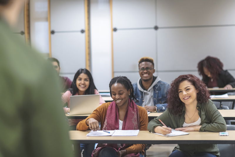 A multi-ethnic group of students sits in a lecture hall listening to their classmates deliver a presentation. They are attentive while smiling and encouraging their mate to continue.