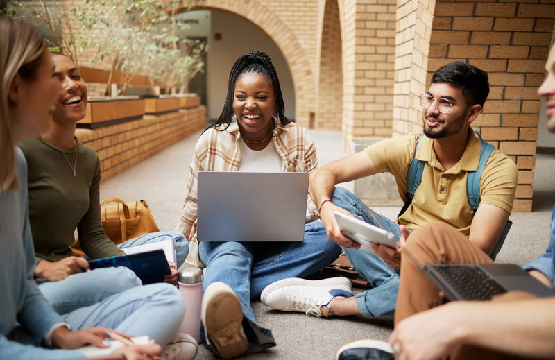 A group of five people sitting in a circle on a floor, using laptops and tablets, smiling and talking in a bright, open indoor space.