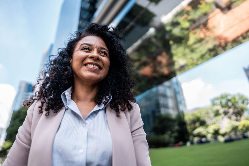 Woman smiling outdoors in a city setting with modern buildings and greenery in the background.