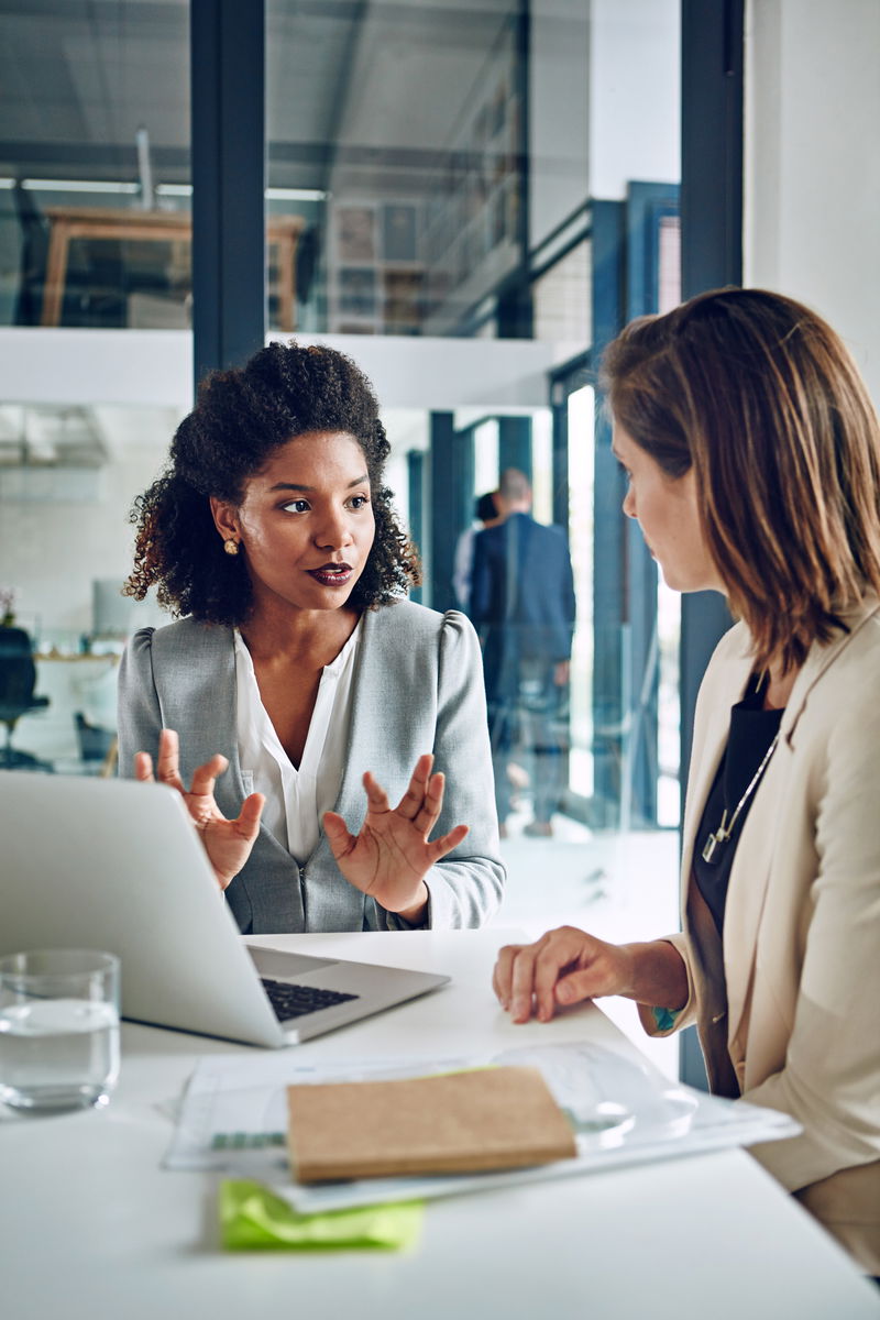 Two women are having a discussion at a desk with a laptop and documents. One woman gestures while speaking, and the other listens attentively. They are in a modern office setting.