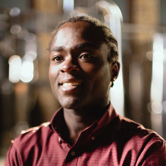 A person wearing a red checkered shirt and a watch stands with arms crossed, smiling, with a blurred background.