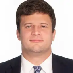 A man in a suit and tie, with short brown hair, looks straight at the camera against a plain white background.