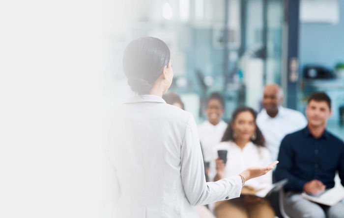 A woman in a light-colored blazer is speaking to a seated group of people in a bright, modern office setting.