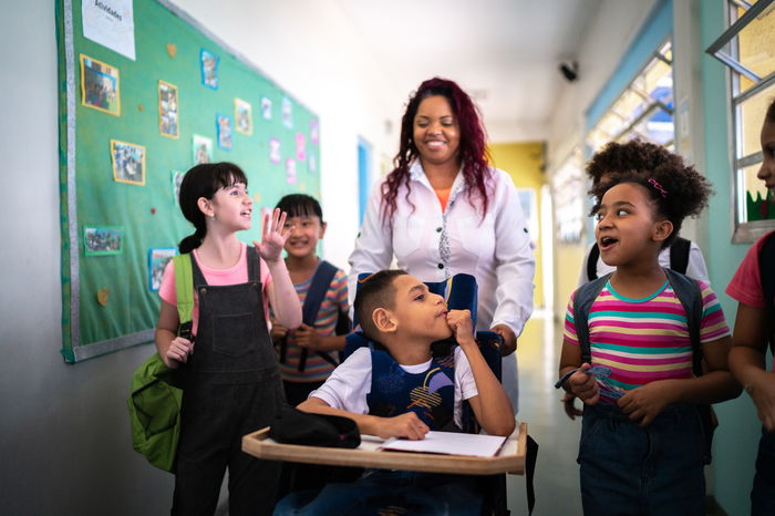 A group of children walk down a school hallway with a teacher. One child is in a wheelchair, accompanied by the teacher, while others chat and smile. A bulletin board is visible on the wall.