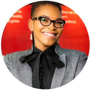 Person wearing glasses and a grey blazer with a black bow tie smiles against a blurred red background.