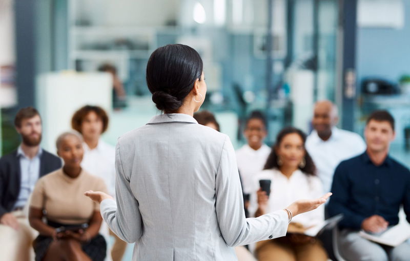 A person in a suit speaks to an attentive audience in a bright conference room.