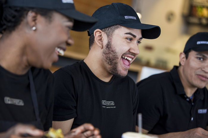 Three people wearing uniforms and caps share a laugh at a table indoors.