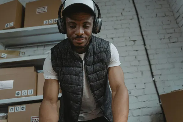 A person wearing headphones and a vest stands in a room with shelves and boxes against a brick wall.