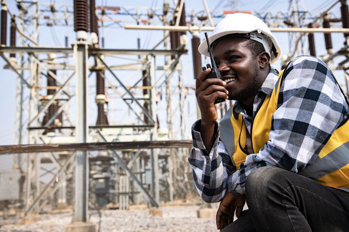 A worker in a hard hat and safety vest uses a walkie-talkie at an electrical substation.