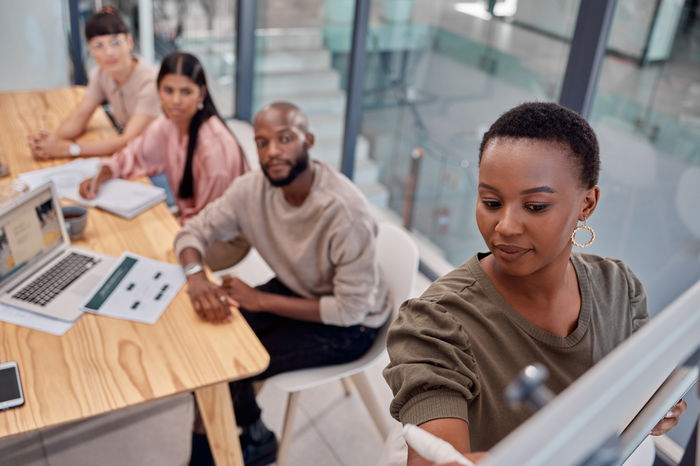 A woman writing on a whiteboard, while four people sit at a conference table, focusing on a presentation.