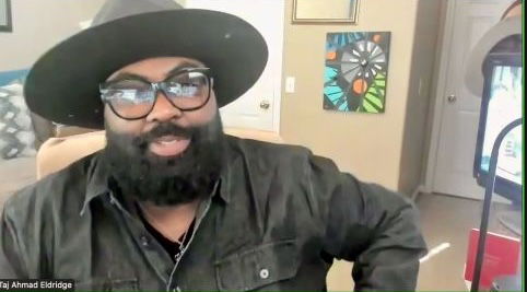 A person with a beard and glasses wears a large-brimmed hat and dark shirt, sitting in a room with colorful artwork on the wall behind them.