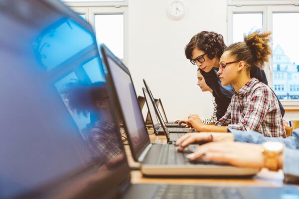 People using laptops in a classroom, with a teacher assisting a student.