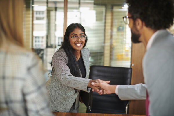 Woman in a gray blazer smiles while shaking hands with a man in an office setting, with another person partially visible in the foreground.