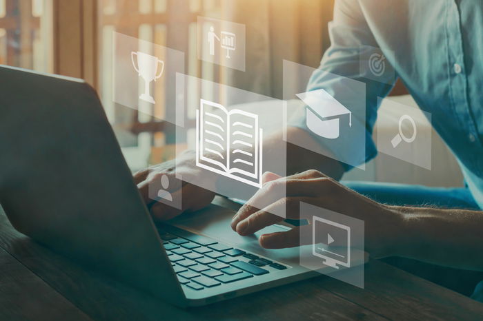 Person typing on a laptop with floating digital icons including a book, trophy, and graduation cap, symbolizing online learning and achievement.