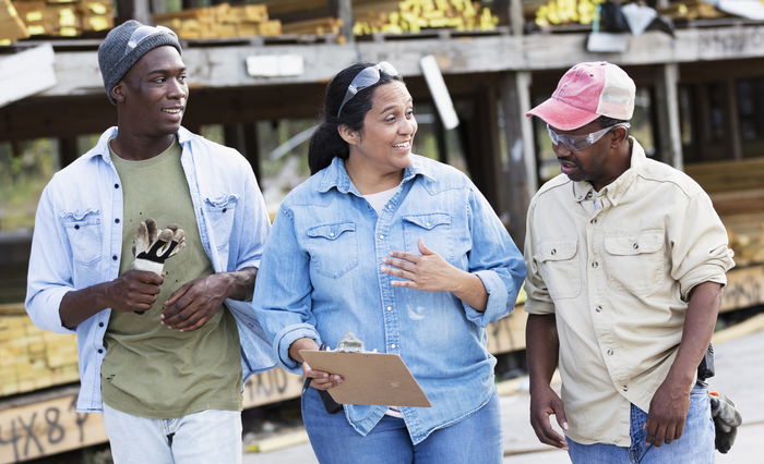 Three construction workers wearing casual work clothes stand outdoors, talking and smiling; one holds gloves, another a clipboard, and lumber is stacked in the background.