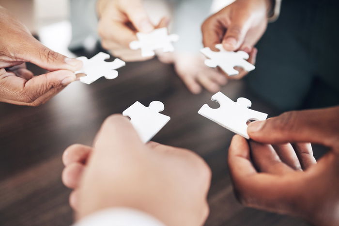 Five people each hold a white puzzle piece over a table, appearing to assemble the pieces together.