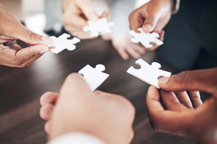 Five people each holding a white puzzle piece, gathering in a circle over a wooden table surface.