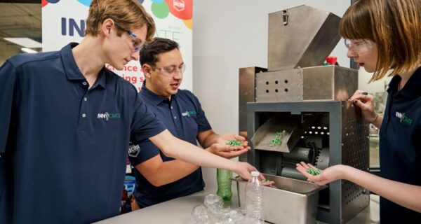 Three individuals demonstrate a machine processing plastic bottles into small green pellets, wearing safety glasses and blue shirts in an industrial setting.