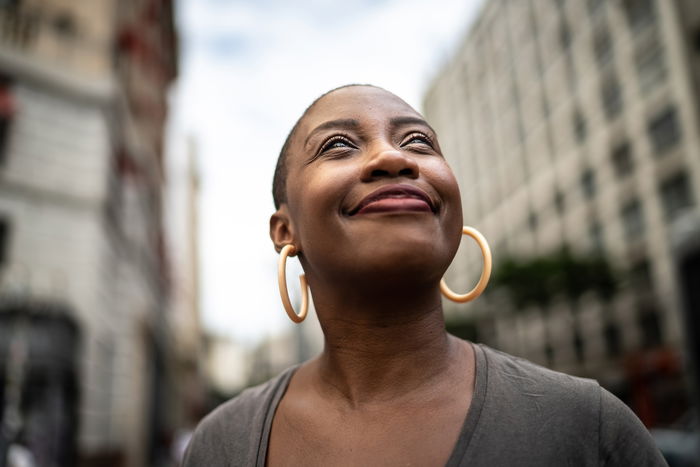 Person with short hair and large hoop earrings smiles while looking upwards. Tall buildings are visible in the blurry background.