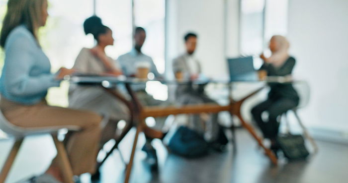 A blurred image of five people sitting around a table in a meeting room, with a laptop and coffee cups visible on the table.