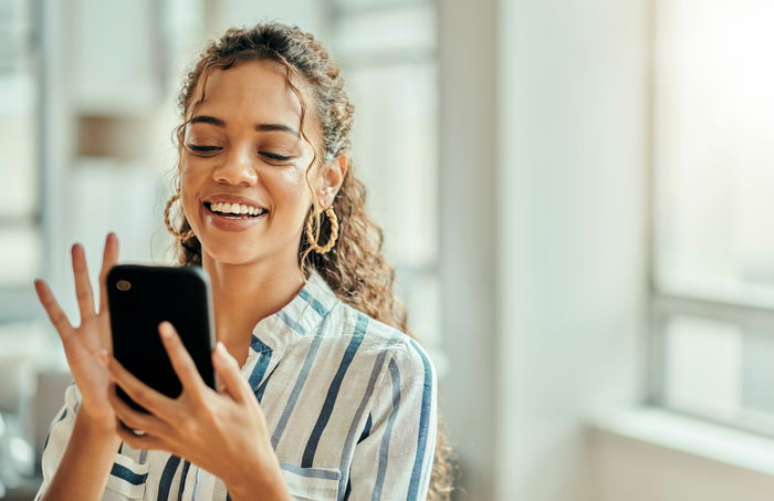 A woman smiles while looking at her smartphone indoors. She is wearing a striped shirt and hoop earrings, with sunlight streaming in through large windows.