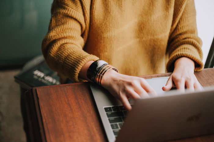 Person wearing a yellow sweater types on a laptop at a wooden desk.