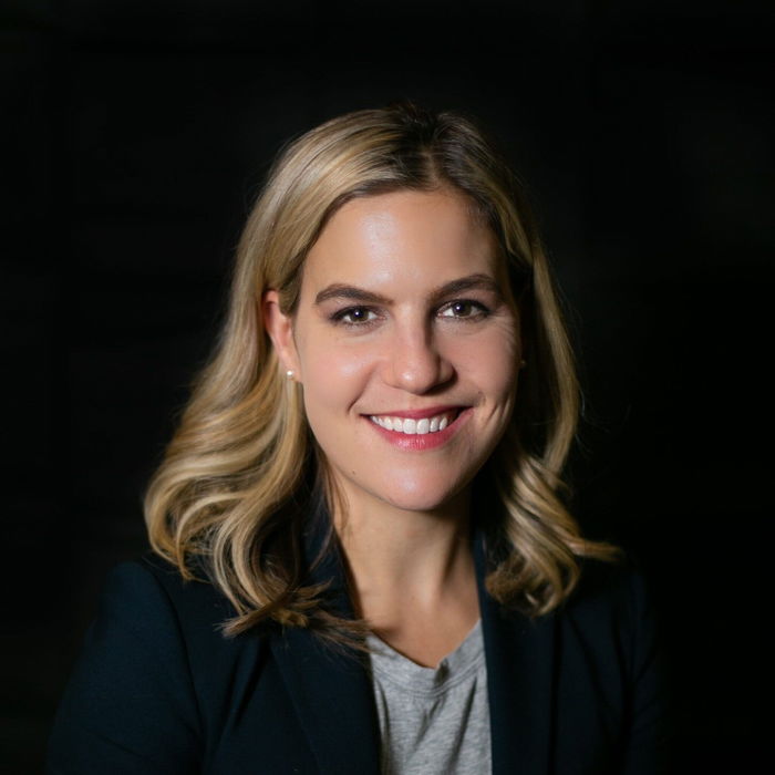 A smiling person with shoulder-length blonde hair wearing a black blazer and gray shirt against a dark background.