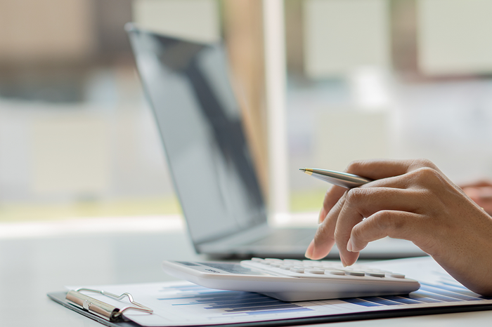 Person using a calculator and holding a pen, with an open laptop in the background on a desk.