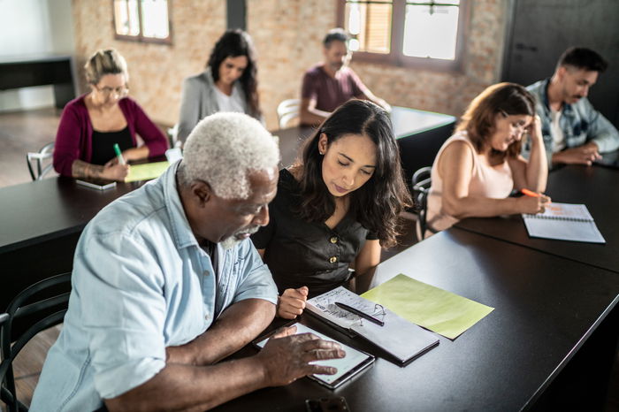 A diverse group of adults seated at desks in a classroom setting, engaged in writing and discussion, with a focus on a man and woman collaborating on a task.